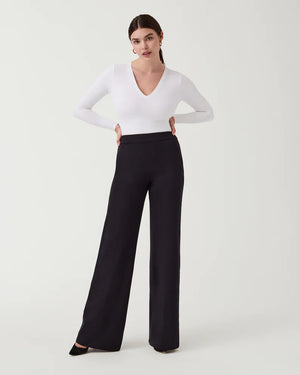 A woman stands against a plain background wearing the SPANX Perfect Pant Wide Leg in black—crafted from premium ponte fabric—paired with a white long-sleeve V-neck top and black heeled shoes.
