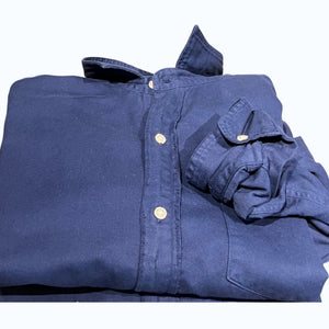 A man with short dark hair and a beard stands sideways with arms crossed, wearing the FRANK & EILEEN LUKE Men's Button Up Shirt in Famous Denim Washed Navy, blue jeans, and white sneakers. He is indoors by a light wall.