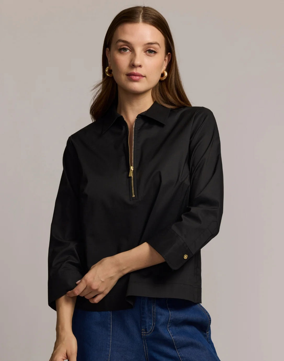 A woman with long brown hair is wearing the HINSON WU - ALESSA 3/4 SLEEVE SHIRT in black, crafted from stretch polished cotton, paired with blue jeans. She stands against a plain light background and looks at the camera with a neutral expression.