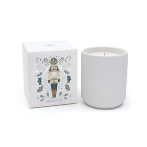 A plain NUTCRACKER SIBERIAN FIR CANDLE in a matte vessel sits next to its square box featuring a nutcracker illustration and holly motifs.