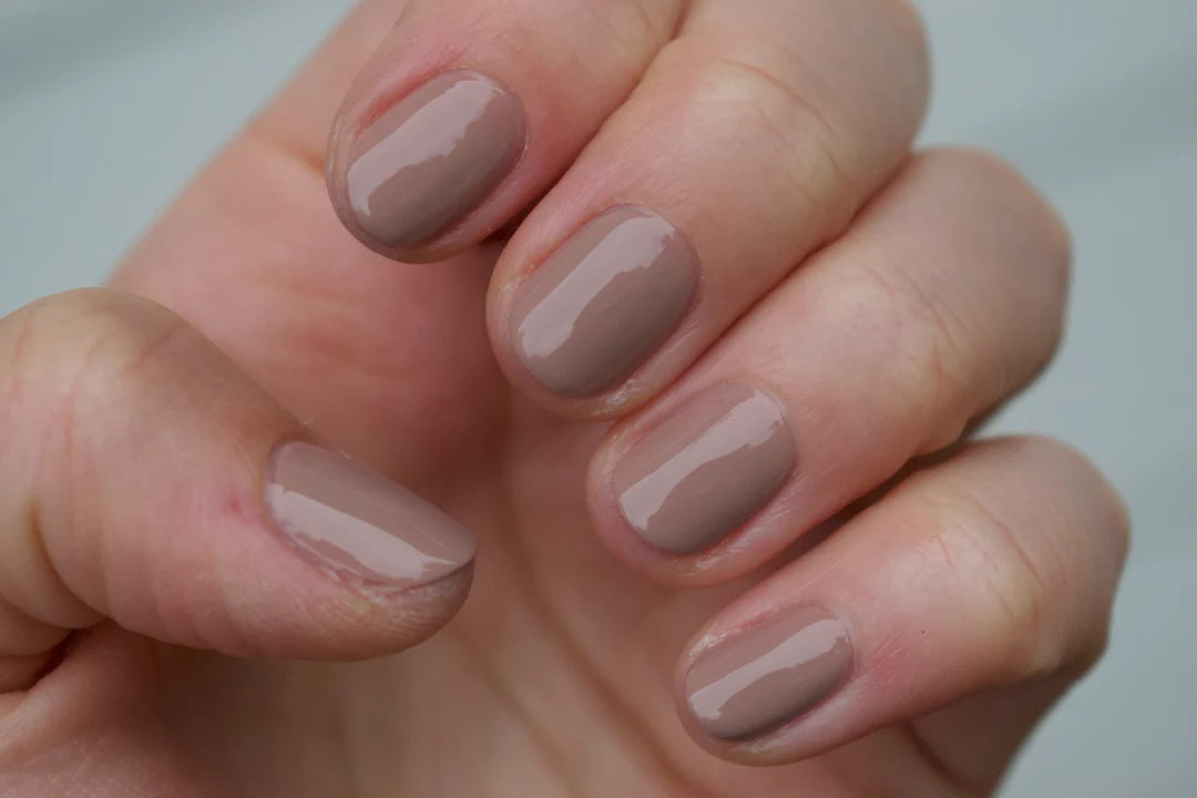 A glass bottle of TENOVERTEN - NAIL POLISH from the brand TEN OVER TEN, featuring a white cap and a dusty pink nail color. The front label displays "TEN OVER TEN" in white uppercase letters. This vegan, eight-free formula ensures both beauty and peace of mind. The background is plain and light-colored.