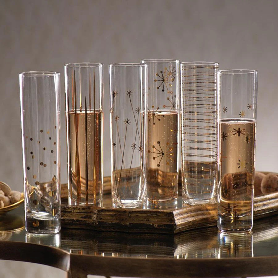 A set of six LA FETE GOLDEN DECAL CHAMPAGNE FLUTES by ZODAX, featuring tall, clear glasses adorned with decorative gold patterns such as stars, lines, and dots, showcased on a reflective surface. For details on dimensions or capacity, feel free to call the store.