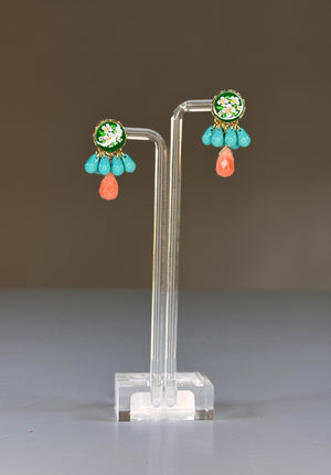The SANDRINE FRANCE STUDIO GRACIE EARRINGS feature colorful floral motifs with green, white, and gold tops, blue beads, and an orange teardrop bead. Handmade in NYC, they are displayed on a clear stand against a plain gray background.