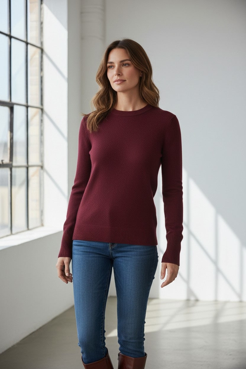 A woman with wavy brown hair, wearing the KINROSS MODERN CREW NECK CASHMERE SWEATER in Cordovan and blue jeans, stands in a bright room filled with natural light from large windows.