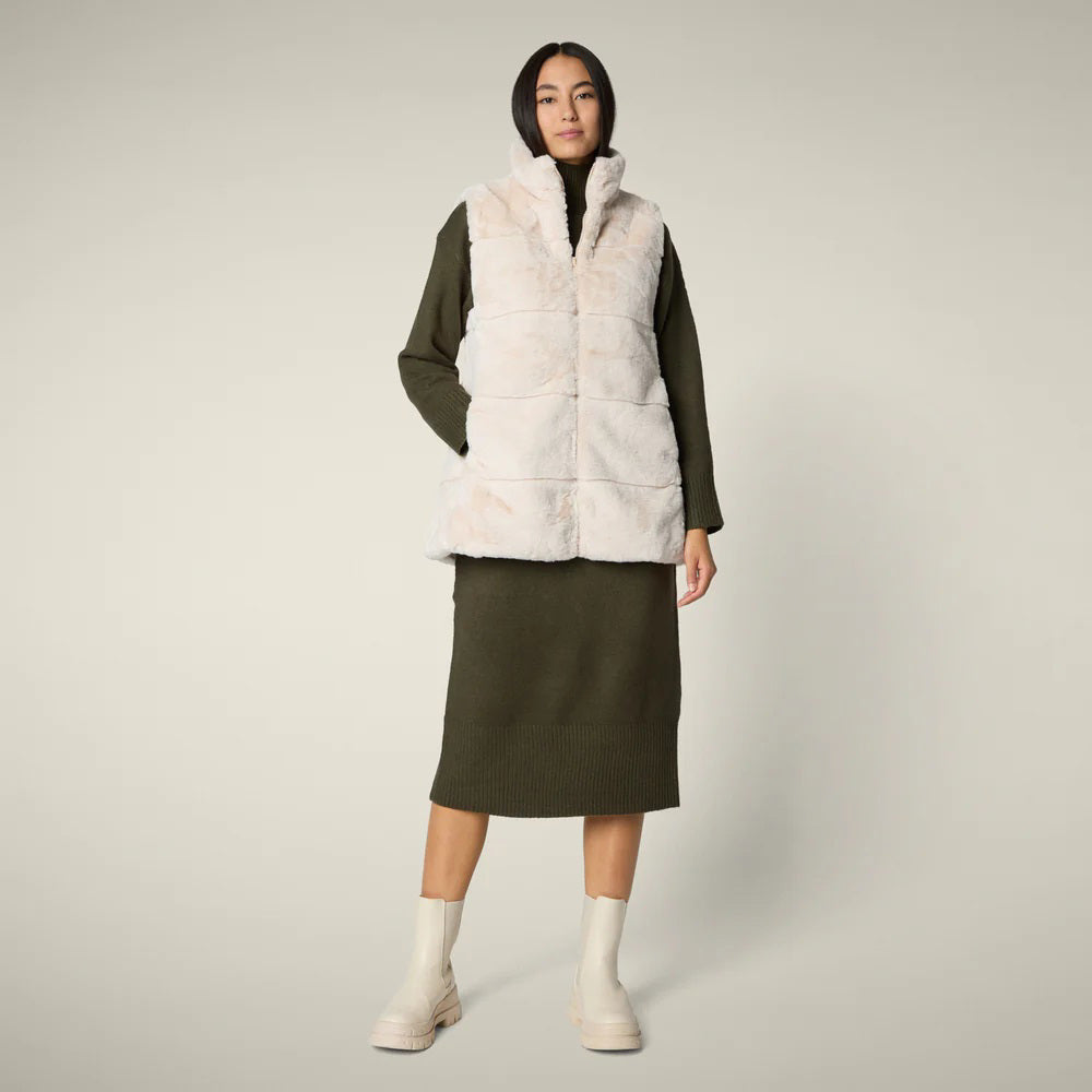 A woman with straight blonde hair wears the SAVE THE DUCK NIVES REVERSIBLE VEST over a white long-sleeve top, beige wide-leg pants, and black shoes, standing against a plain background and highlighting the vest&#39;s chic style.