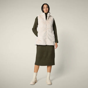 A woman with straight blonde hair wears the SAVE THE DUCK NIVES REVERSIBLE VEST over a white long-sleeve top, beige wide-leg pants, and black shoes, standing against a plain background and highlighting the vest's chic style.