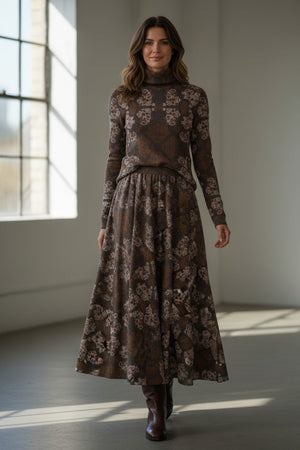 A person wearing the FRANCO FERRARI - FRAGOLINA SKIRT, paired with a matching long-sleeve top and brown boots, stands indoors near a sunlit window.