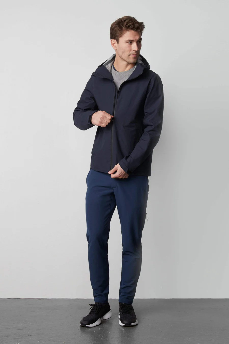 A man stands in front of a white wall wearing the MACK WELDON STORMCHASER JACKET with adjustable hood, paired with a gray shirt, blue pants, and black sneakers. He looks to the side while holding the jacket's zipper.