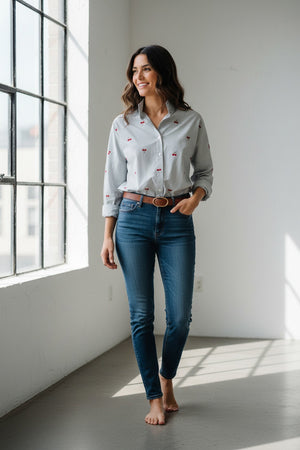 A woman with blonde hair in a ponytail, dressed in the MERSEA Emerson Button Up Shirt in Cherries On Top by MERSEA and blue jeans, holds a red clutch while standing outdoors near green trees and patterned tiles, smiling and looking to the side.