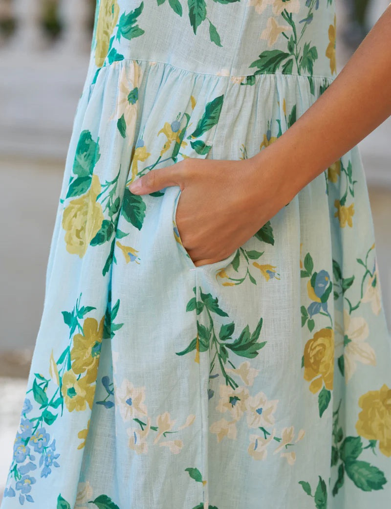 A woman with blonde hair stands outside on a stone patio in the FRANK &amp; EILEEN Daphne Popover Dress in Italian Linen Summer Scatter, paired with brown sandals. Greenery and a large planter are visible in the background.