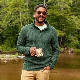 Wearing the CRIQUET Quilted Pullover in Heather Ivy, a person stands by a stone wall in khaki pants, holding a patterned mug and smiling, with greenery in the background—highlighting this classic-fit all-season layer from CRIQUET.
