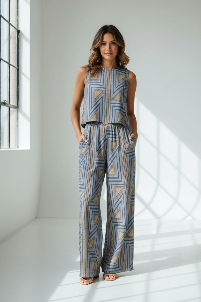 A woman stands against a plain white background wearing the BRITON COURT SARA PANT by BRITON COURT, a sleeveless jumpsuit with wide-leg pants featuring a blue and tan geometric pattern.
