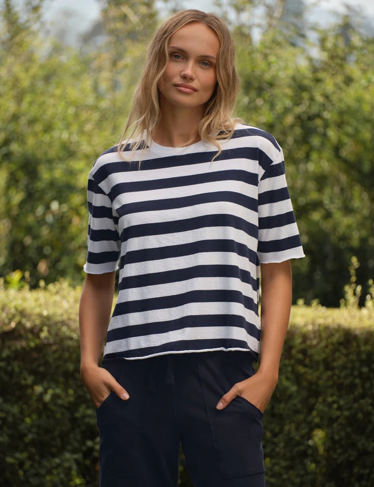 A person stands outdoors amidst green bushes and trees on a sunny day, wearing the FRANK & EILEEN Perry NYC Crewneck Tee in Vintage Jersey Nautical Stripe—a navy and white striped short-sleeve shirt crafted from 100% cotton—paired with dark pants.