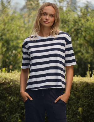 A person stands outdoors amidst green bushes and trees on a sunny day, wearing the FRANK & EILEEN Perry NYC Crewneck Tee in Vintage Jersey Nautical Stripe—a navy and white striped short-sleeve shirt crafted from 100% cotton—paired with dark pants.