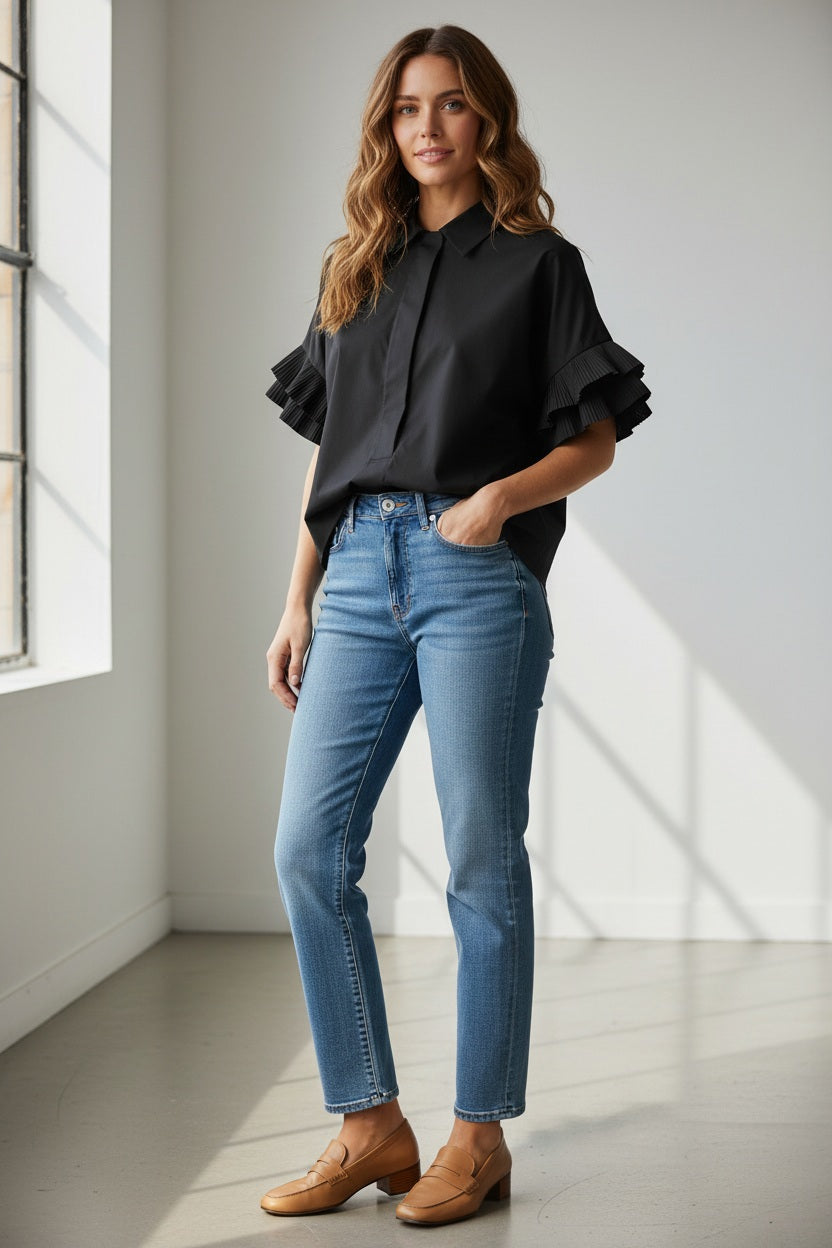 A woman stands indoors near a window, smiling softly in the MEIMEIJ - COTTON SHIRT IN BLACK by MEIMEIJ, featuring ruffled short sleeves. She pairs the top with light blue jeans and tan loafers, her long wavy hair complementing the relaxed silhouette.