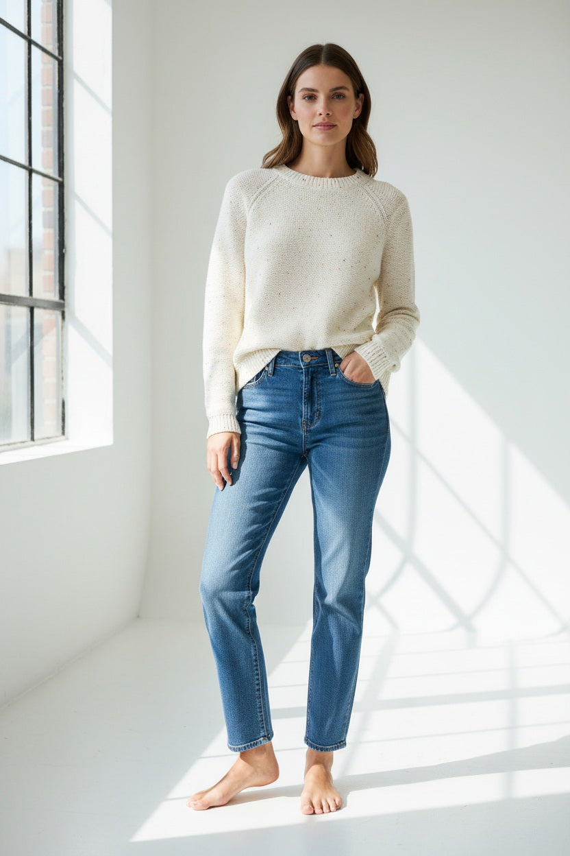 A woman with long brown hair wears the HINSON WU - QUEENIE Long Sleeve Sequin Sweater in Pearl White and blue jeans, standing against a plain light background and looking slightly to the side with a relaxed expression.