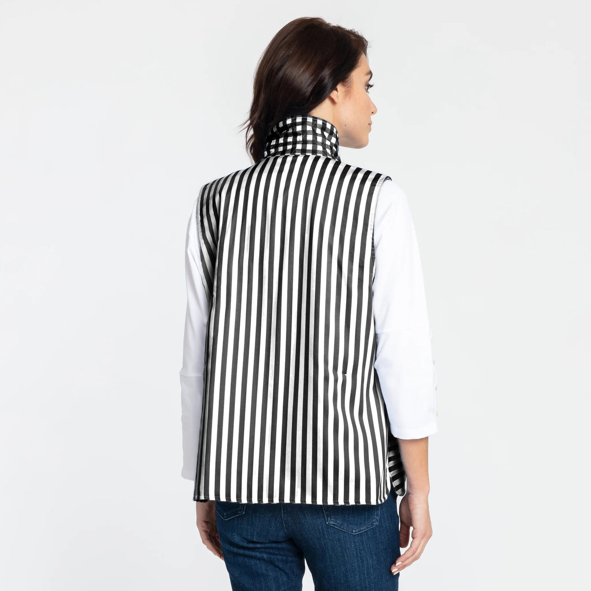 A woman with shoulder-length dark hair is wearing the HINSON WU - LAUREN VEST, which is fully reversible and features a blue and white gingham pattern, over a white long-sleeve shirt. She is standing sideways, looking at the camera with her left hand resting near her waist. The background is plain white.