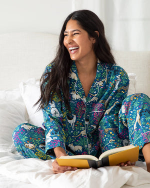 A woman in PRINTFRESH's colorful "Must Love Dogs" organic cotton pajama set stands barefoot on a light rug against a white paneled wall. Her dark hair falls freely as she casually places her hands in her pockets, facing slightly to the side.