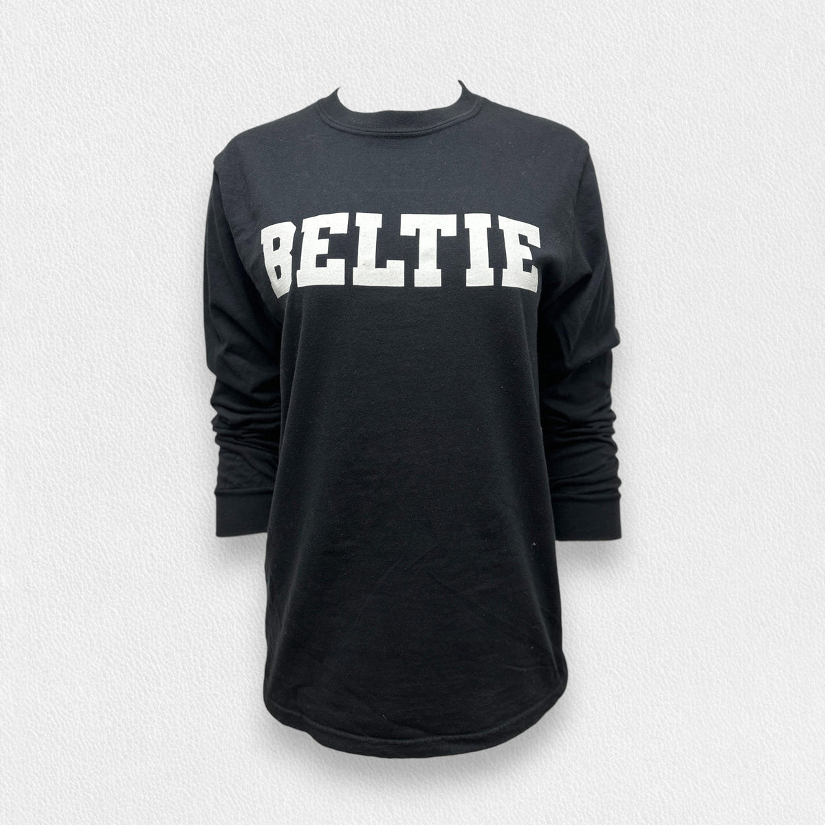 A black long sleeve tee shirt from the Fearrington Lifestyle Collection by Comfort Colors, crafted from premium fabric with the word &quot;BELTIE&quot; printed in large, bold, white capital letters on the front, neatly folded and laid on a white textured surface.