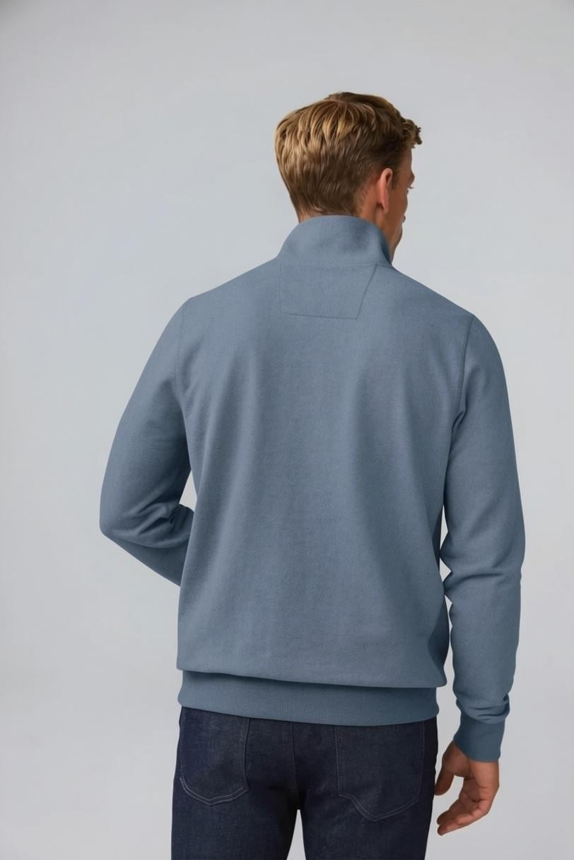 A man with short light brown hair stands in profile, wearing the MACK WELDON Ace 1/2 Zip Sweater in Herringbone and dark pants, one hand in his pocket, against a plain light gray background.