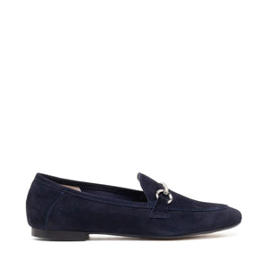 The MICHELE LOPRIORE - LEYLA LOAFER is a navy blue suede loafer adorned with a silver horsebit ornament on the vamp. Made in Italy and crafted from genuine leather, this shoe features a low heel and boasts a sleek, streamlined silhouette against a plain, off-white textured surface.