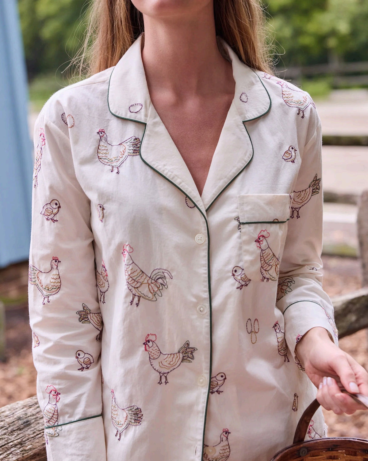 A woman dressed in the PRINTFRESH Embroidered Hens Long Pajama Set in Buff smiles as she stands by a wooden fence, holding a basket and watching chickens peck at the ground in a farmyard setting, with trees and a path visible behind her.