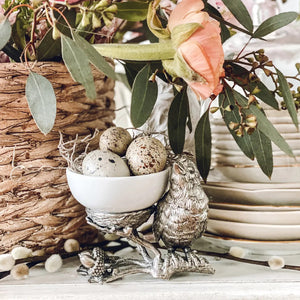 A small white bowl of butter sits on Vagabond House's Songbird Salt Cellar, showcasing intricate pewter designs with delicate feathers. The scene is set on a dark surface with blurred greenery in the background.