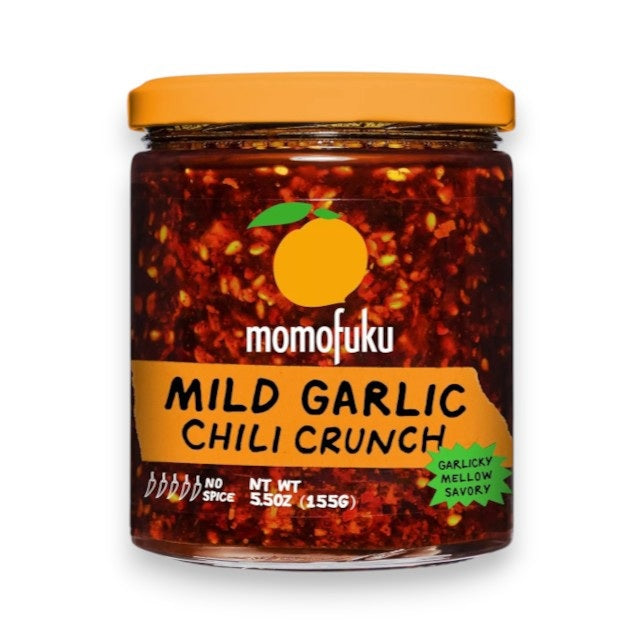 A jar of MOMOFUKU - GARLIC CRUNCH, the ultimate crunchy garlic condiment from MOMOFUKU, with an orange lid. The label displays an eye-catching orange graphic and emphasizes “Garlic Crunch.” Inside, the vibrant red mixture is speckled with visible chili flakes, highlighting its signature crunchy texture.
