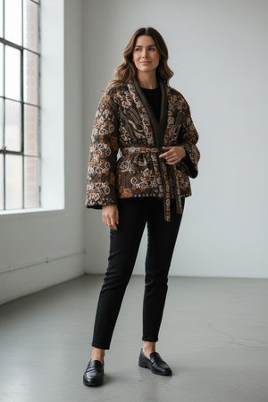 A woman stands indoors by a window, smiling softly with wavy hair. She wears the FRANCO FERRARI - REVERSIBLE KIMONO JACKET, which features a patterned velvet design and a subtle kimono silhouette, paired with a black top, black pants, and black loafers.