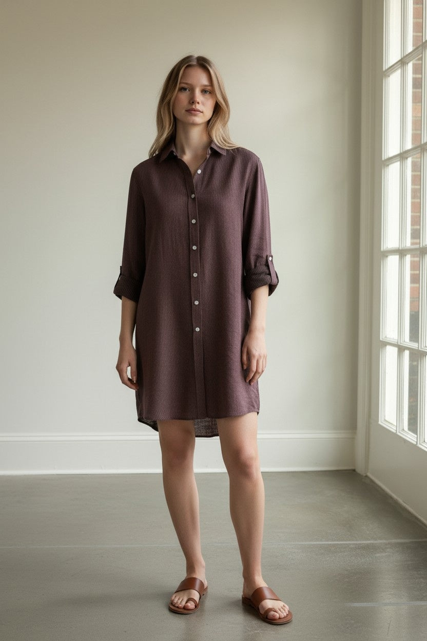 A woman stands indoors near a large window bathed in natural light, wearing the EMILY PHILLIPS - THE MINI SHIRTDRESS IN BORDEAUX STRIPE LINEN with rolled-up sleeves and brown sandals.