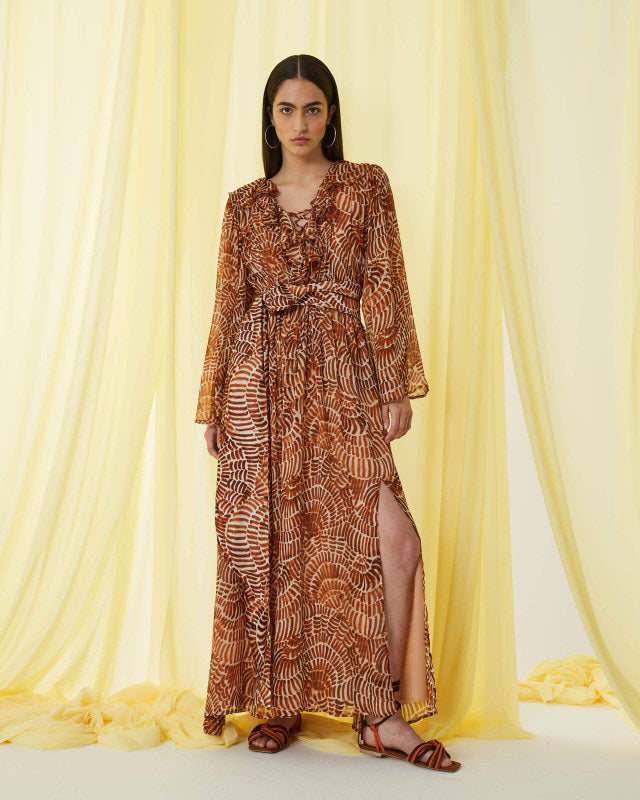 The SFIZIO - RUST PRINT RUFFLED DRESS by SFIZIO is a long, full-sleeved, flowy dress made in Italy, featuring a brown and beige spiral pattern. It boasts a V-neckline, ruffled detailing on the bodice, and an elastic waistband. Crafted from lightweight viscose creponne, it’s perfect for warm weather.