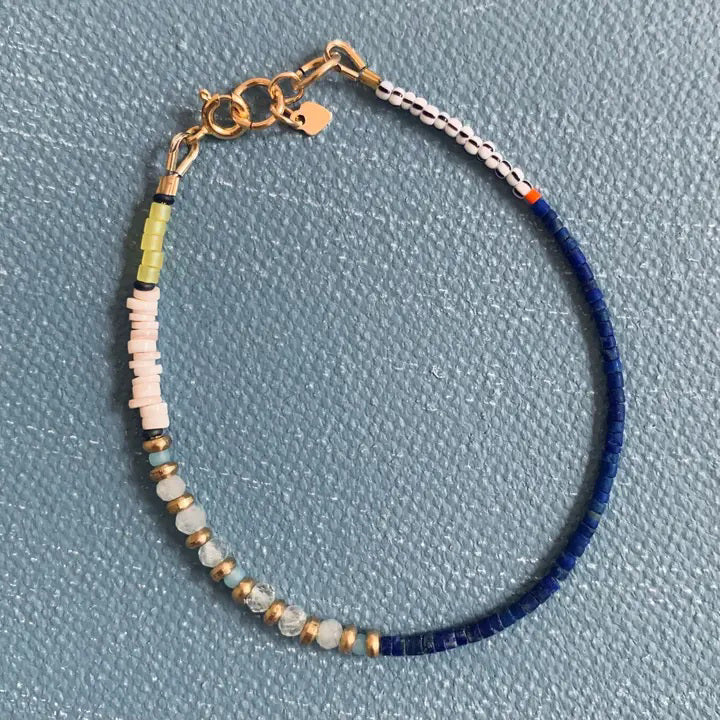 The ALICE RISE - AQUAMARINE BRACELET by ALICE RISE features blue, white, green, and light pink beads with aquamarine stone accents, a gold clasp, and handmade North Carolina craftsmanship. Patterned beads rest on a textured blue surface.