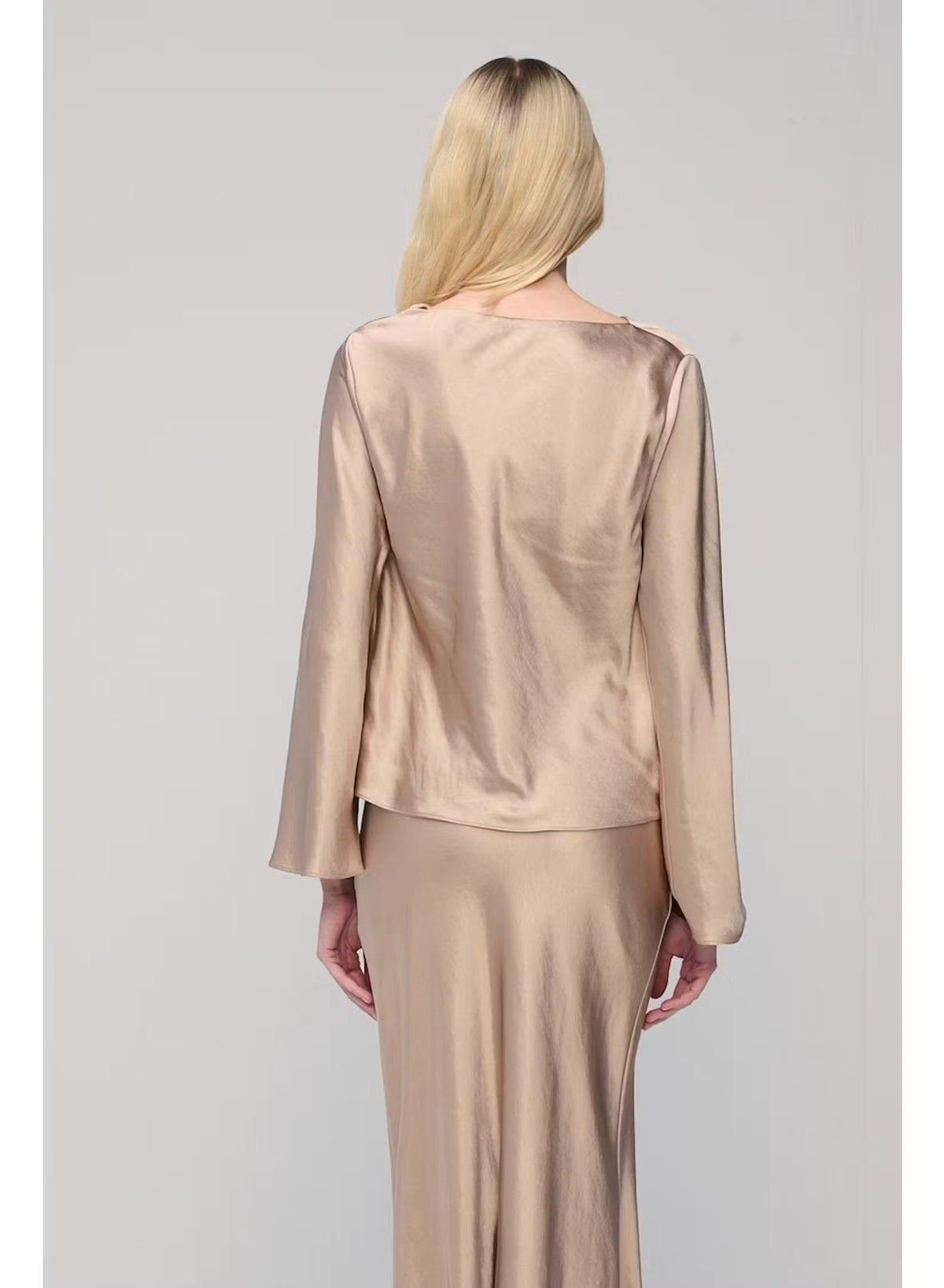 A woman with long blonde hair poses against a plain light gray background, wearing the MILVA SATIN BLOUSE IN SAND by MILVA—a shiny, champagne-beige long-sleeve blouse featuring a chic cowl neckline—paired with a matching skirt and accessorized with bracelets on her left wrist.