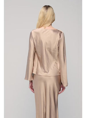 A woman with long blonde hair poses against a plain light gray background, wearing the MILVA SATIN BLOUSE IN SAND by MILVA—a shiny, champagne-beige long-sleeve blouse featuring a chic cowl neckline—paired with a matching skirt and accessorized with bracelets on her left wrist.