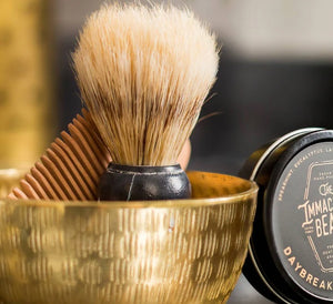 THE IMMACULATE BEARD - SHAVE BRUSH by THE IMMACULATE BEARD, featuring a black handle and faux boar bristles, stands upright on a wooden tray with a green plant and white tiled wall blurred in the background.