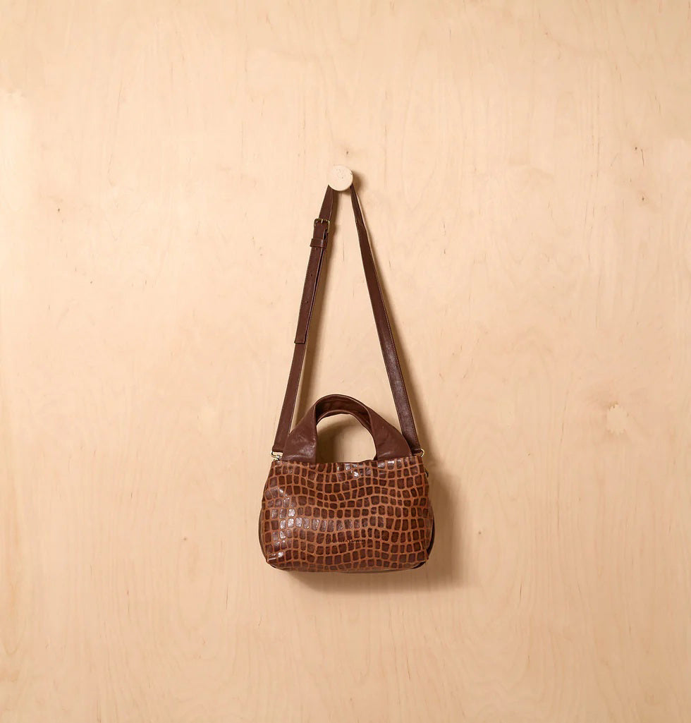 The DANIELLA LEHAVI EVELINE Satchel in Brown Mosaic, featuring double handles, a croco-inspired texture, and an adjustable shoulder strap, hangs on a wall hook against a light wooden background.