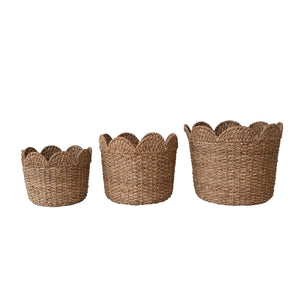 Three handwoven CREATIVE COOP BRAIDED BANKUAN & RATTAN BASKETS are displayed against a light-colored wall. The largest basket contains decorative dried grasses, the medium basket holds patterned pillows, and the smallest basket has rolled-up fabrics with various prints and colors. These eco-friendly pieces add charm to any room.