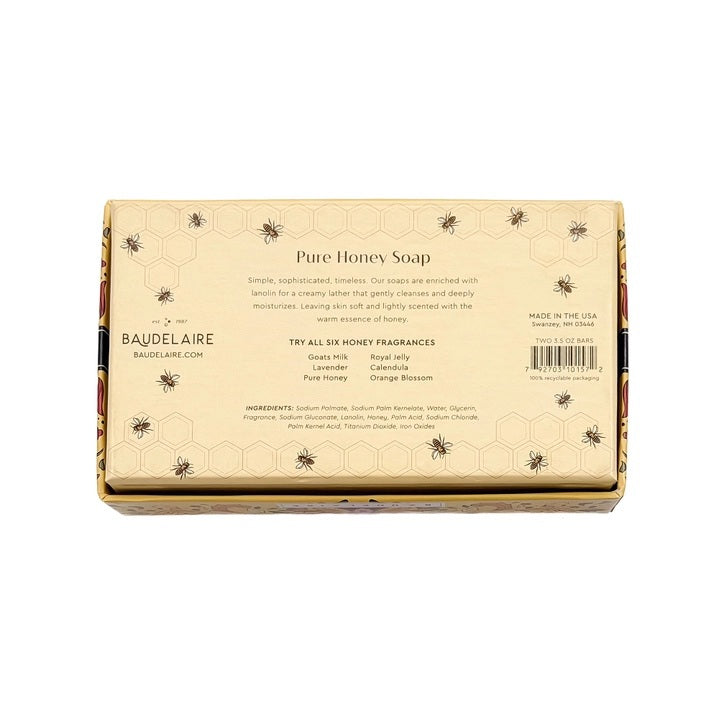 A floral box labeled “BAUDELAIRE - PURE HONEY SOAP 3.5 OZ, 2 BAR GIFT BOX” sits beside two honeycomb-shaped BAUDELAIRE Pure Honey Soaps featuring a bee design—each a gentle, triple-milled soap ideal for sensitive skin—accompanied by a white tray, yellow flowers, and stones on a white surface.