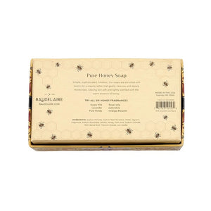 A floral box labeled “BAUDELAIRE - PURE HONEY SOAP 3.5 OZ, 2 BAR GIFT BOX” sits beside two honeycomb-shaped BAUDELAIRE Pure Honey Soaps featuring a bee design—each a gentle, triple-milled soap ideal for sensitive skin—accompanied by a white tray, yellow flowers, and stones on a white surface.