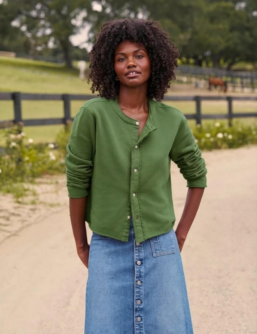 A woman stands outdoors on a dirt path, wearing the FRANK & EILEEN JACKIE Sweatshirt Cardigan in Triple Fleece Nori from the California Heritage Collection—a green button-up sweater paired with a blue denim skirt. There is a wooden fence, green grass, and trees in the background.