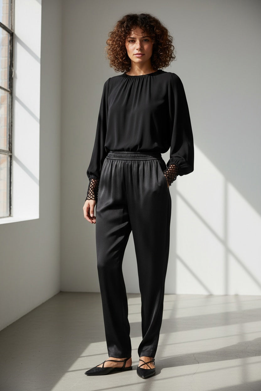 A woman with short dark hair faces away, wearing the MILVA MI - CROCHET CUFF BLOUSE by MILVA, black pants, and pointed black shoes. She holds a silver textured clutch against a plain gray background.