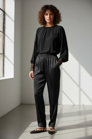 A woman with short dark hair faces away, wearing the MILVA MI - CROCHET CUFF BLOUSE by MILVA, black pants, and pointed black shoes. She holds a silver textured clutch against a plain gray background.