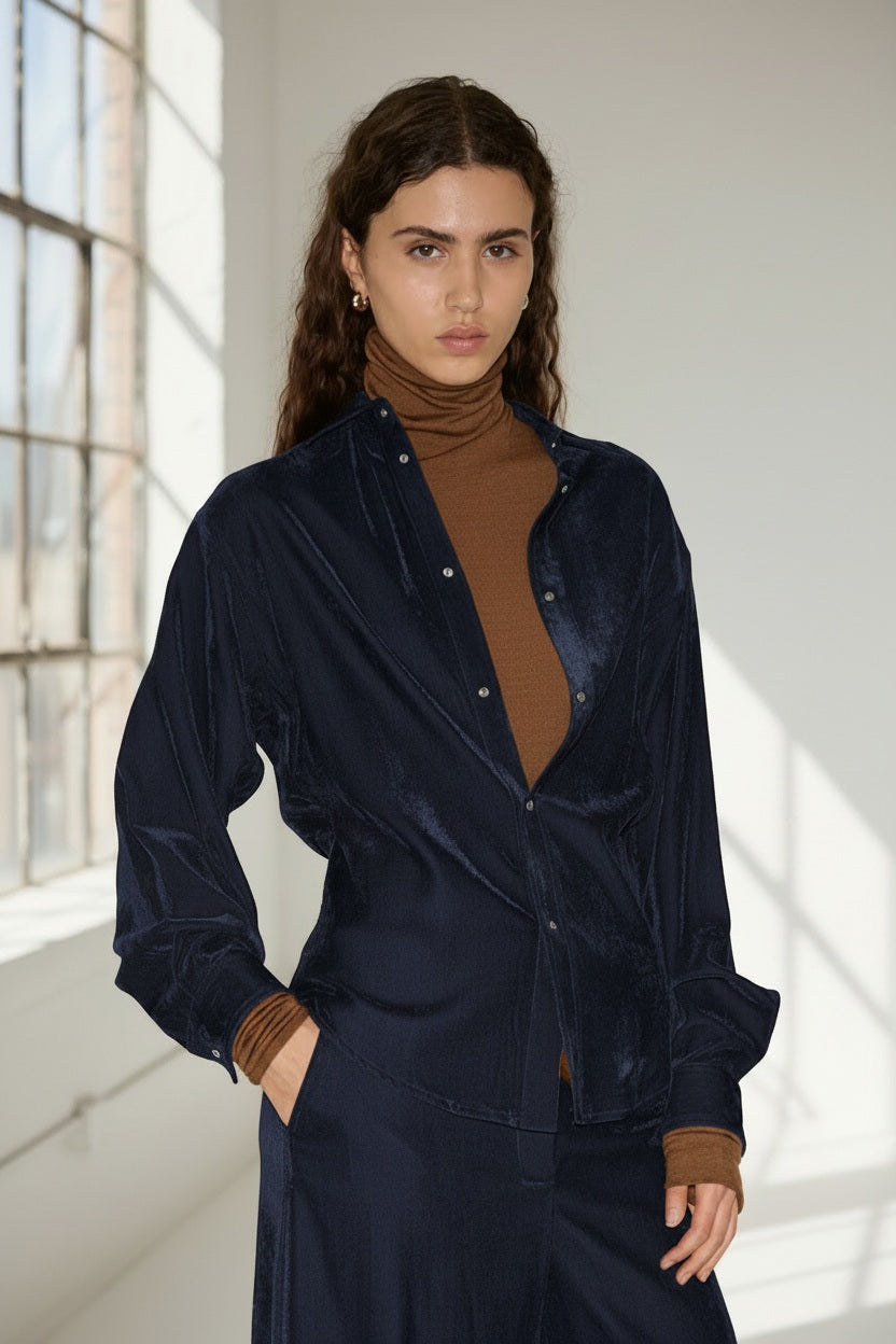 Wearing the NUDE Fitted Shirt in Blue by NUDE, a woman with long curly hair layers it over a brown turtleneck and pairs it with matching pants. She stands indoors near large windows with natural light, epitomizing relaxed elegance in this luxurious Italian viscose blend ensemble.