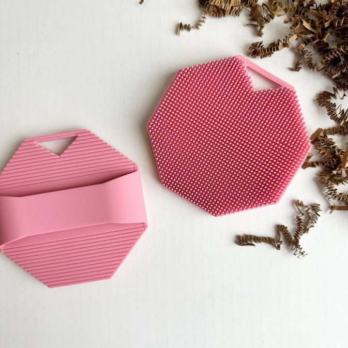 A rose-colored, octagonal SILICONE BODY AND EXFOLIATION BRUSH by SMILE BOUTIQUES, featuring raised dots for gentle exfoliation, rests on a white surface. Brown paper packing material is scattered to the right of the brush.