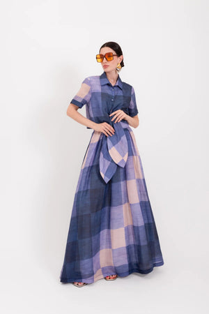 A person stands facing away, wearing the LAVI - MENTA MAXI SKIRT IN QUADRI by LAVI, designed in Italy. The maxi skirt features a fitted waist and a long, flowing silhouette with a blue and beige large checkered pattern reminiscent of oversized plaid. The background is plain white.