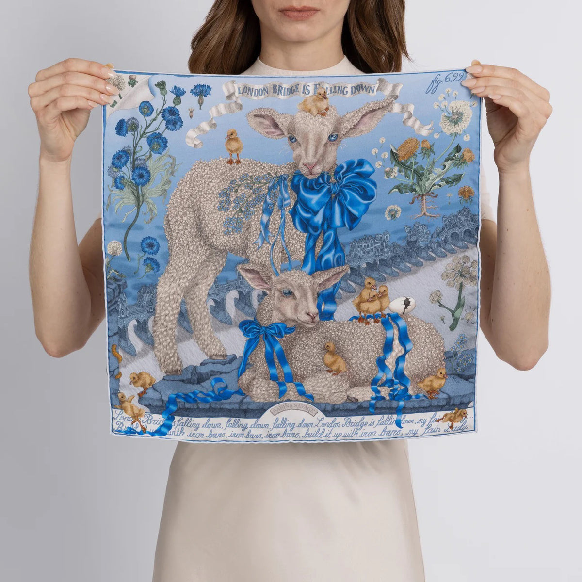 The SABINA SAVAGE - LONDON BRIDGE IS FALLING DOWN SILK TWILL SCARF 42CM POWDER/FROST from SABINA SAVAGE ENGLAND is artfully folded into a floral shape, showcasing a blue and white pattern with flowers, baby lambs, and intricate details bordered by a bold blue.