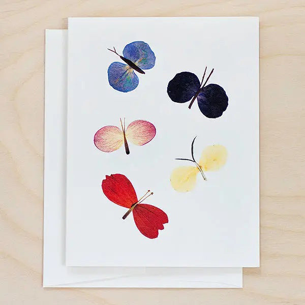 The PETAL PEOPLE PRESS - BUTTERFLY CARD by PETAL PEOPLE PRESS features five vibrant butterflies crafted from petals on a white background, accompanied by green leaves and office supplies—a charming botanical greeting card for any occasion.