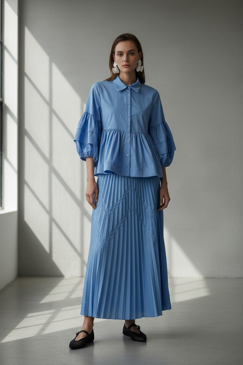 A woman models the MILVA MI Cotton Pleated Skirt in Azzurro Blue by MILVA, paired with a matching flowy top and voluminous sleeves, styled with bold white earrings against a plain gray background.