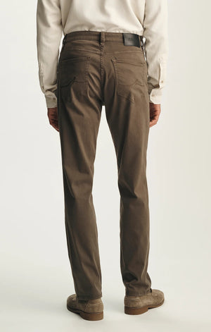 A person is seen from behind, standing against a plain light background, wearing the 34 HERITAGE - CHARISMA RELAXED STRAIGHT PANTS IN HARDWOOD TWILL by 34 HERITAGE. The brown comfort stretch twill pants are paired with a cream long-sleeve shirt and brown shoes.