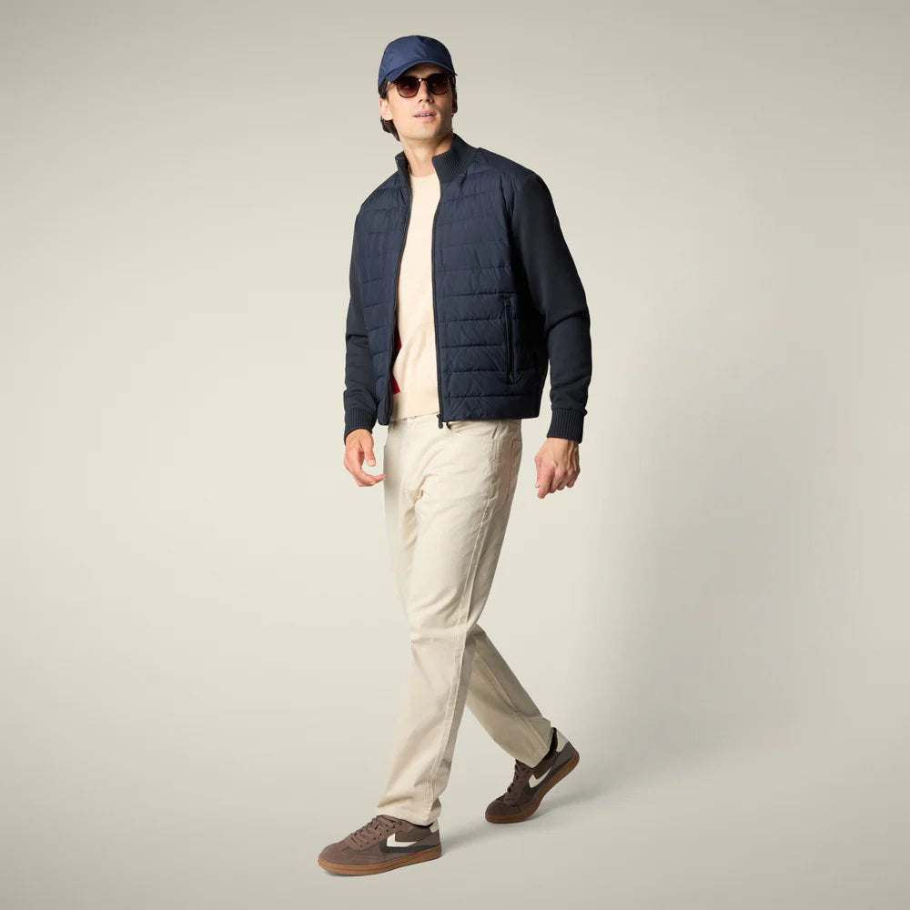 A man with dark hair faces away from the camera, wearing the SAVE THE DUCK - SEDUM JACKET in dark navy by SAVE THE DUCK, paired with light beige pants and brown shoes, set against a plain light background.